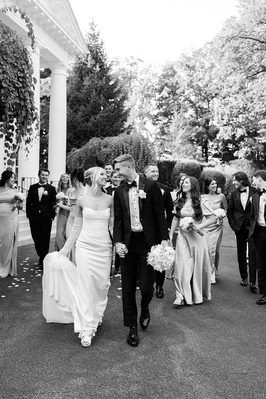 Black and white photo of the couple walking and laughing together, followed by their wedding party.